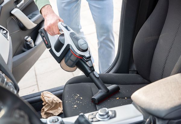 A man is  vacuuming crumbs of a car seat with a soft brush on a cordless unlimited vacuum cleaner.
