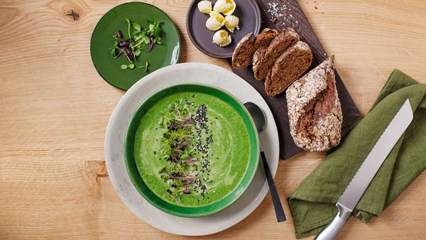 Różne talerze z kremową zieloną zupą, świeże zioła, świeżo pokrojona bagietka z pełnego ziarna i masło.