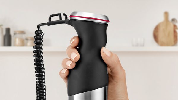 A hand holding the ergonomic handle of a MaxoMixx hand blender with SoftTouch.