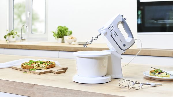 ErgoMixx hand mixer with a mixing bowl and stand next to muffins and pizza.
