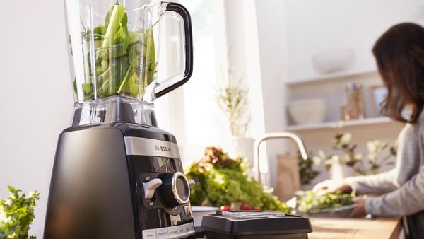  Un robot multifonction rempli de légumes verts, une personne lave une salade en arrière-plan.
