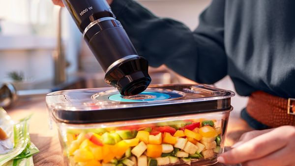 A vacuum container full of fresh vegetables being sealed with a hand blender.