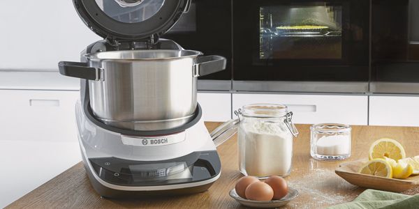 Bosch-Cookit mit offenem Deckel auf einer Arbeitsfläche neben Eiern, Mehl und anderen Zutaten.