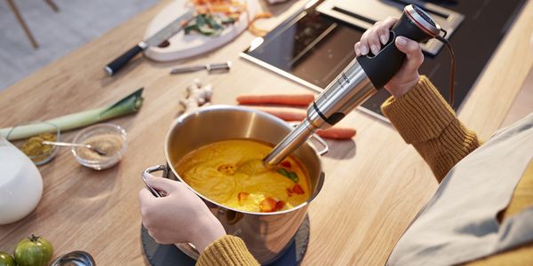 A woman pureeing creamy orange soup with a Bosch hand blender.