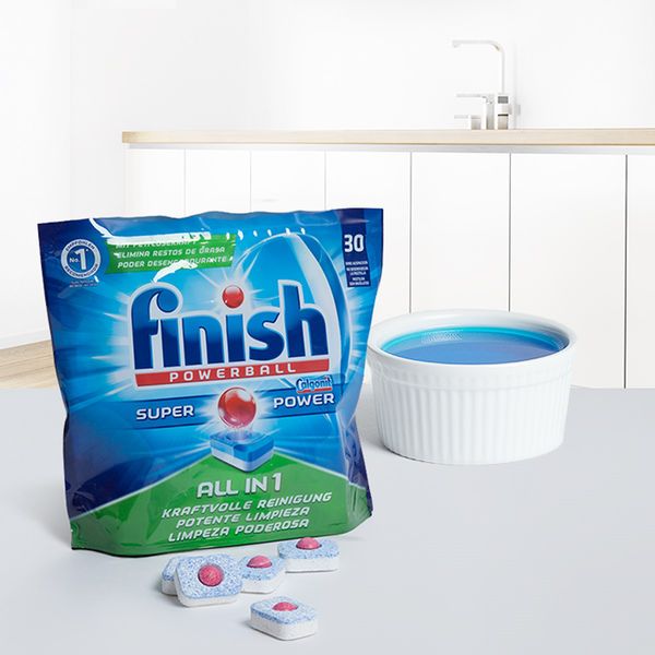 Un ramequin rempli de liquide de rinçage bleu sur un plan de travail dans une cuisine lumineuse, à côté d’un paquet de pastilles pour lave-vaisselle.