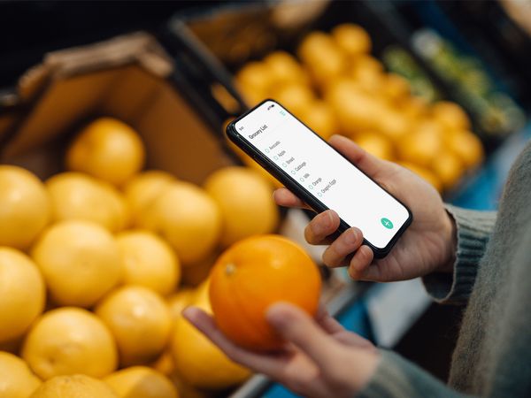 Donna al supermercato con un’arancia in una mano e nell’altra mano lo smartphone per controllare la lista della spesa