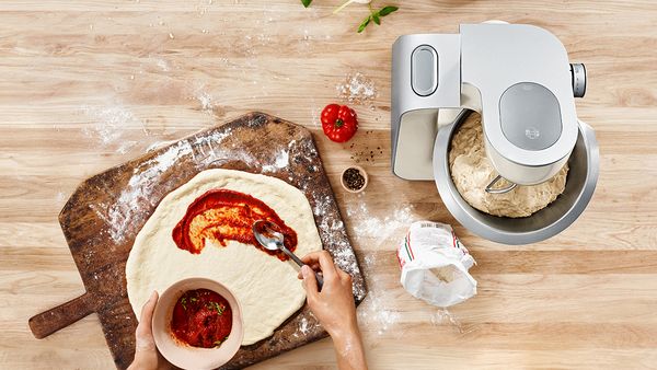 En person brer saus på nyeltet pizzadeig, ved siden av en kjøkkenmaskin.