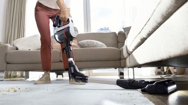 Eine Person reinigt den Boden unter einem Sofa mit dem Bosch Unlimited Akku-Staubsauger und einem knickbaren Staubsaugerrohr.