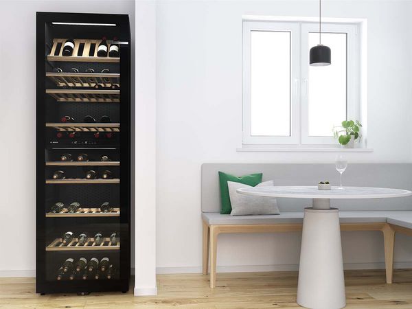 Didelė laisvai pastatoma vyno spinta su keliais vyno buteliais ant medinių lentynų šalia modernios virtuvės kampelio.