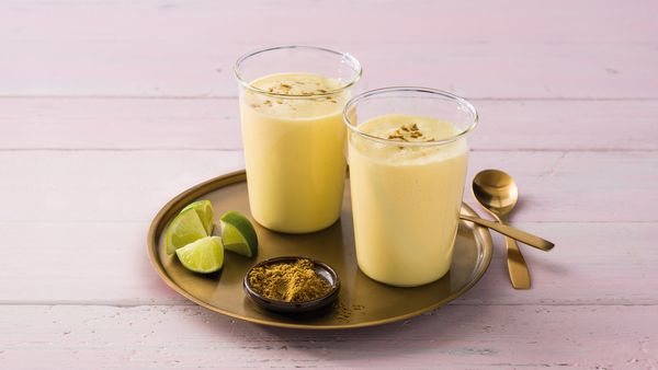 Două smoothie-uri galbene în pahare aranjate împreună cu limete și diferite condimente pe o tavă.