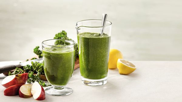 Green smoothie in a glass arranged together with pieces of kale, apple slices and lemons on a table.