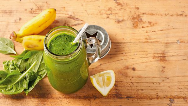 Green smoothie in a glass arranged together with spinach leaves, bananas and a lemon slice on a table.