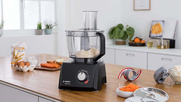 Robot de bucătărie Bosch tip food preocessor lângă o serie de instrumente de bucătărie și ingrediente.