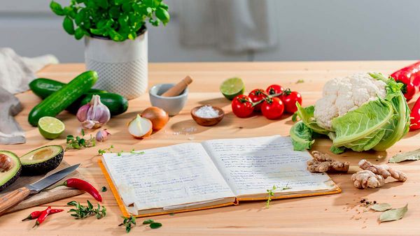 A journal filled with handwritten recipes and surrounded by fresh foods on a wooden countertop.