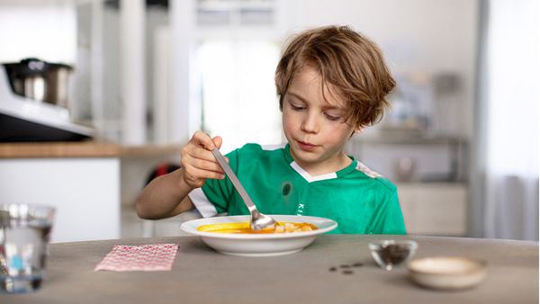 Ein kleiner Junge isst mit Freude eine hausgemachte Suppe, mit dem Cookit im Hintergrund.