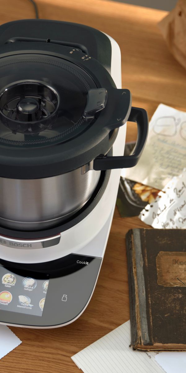 Il robot da cucina Cookit circondato da ricette scritte a mano, con accanto un ricettario dall’aria molto antica.