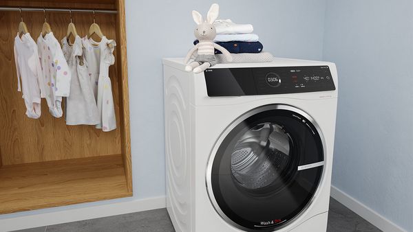 En kosedyrkanin sitter på toppen av et kombinert vask og tørk-apparat på et lyst vaskerom ved siden av et åpent garderobeskap med barnetøy.