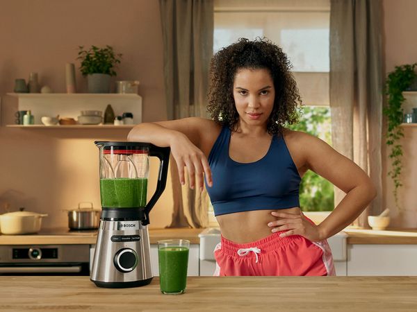 Blender VitaPower Series 4 with green smoothie on the worktop, sporty woman leans casually on blender.