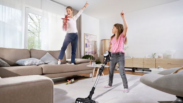 Ein Mädchen steht mit einem Unlimited Akku-Staubsauger in einem Wohnzimmer. Eine Frau steht auf dem Sofa und hält ein Stofftier in der Hand.