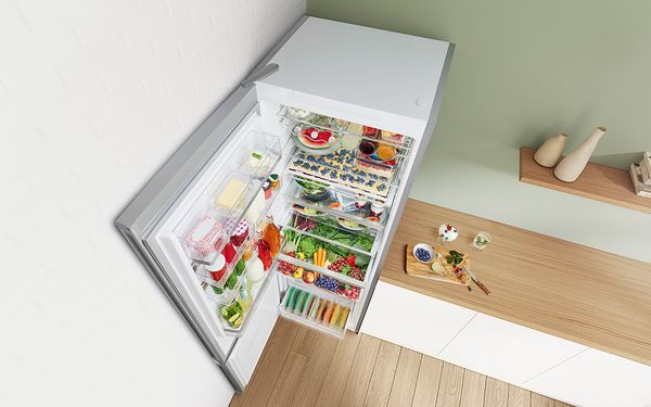 Grand réfrigérateur combiné pose-libre ouvert placé dans un coin de la pièce. La porte du réfrigérateur de 70 cm est ouverte à 90° et est remplie de provisions.