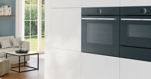 Una cocina moderna y elegante con dos hornos empotrados Bosch sobre un fondo de armarios blancos, con una acogedora zona de asientos a la vista junto a ellos.