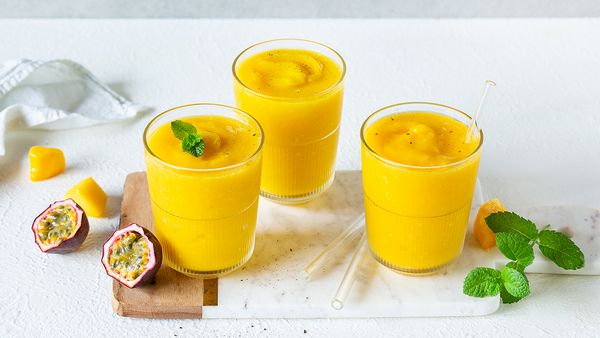 Tres vasos llenos de lassi de mango amarillo brillante se colocan sobre una tabla de madera. A su alrededor se colocan unos trozos de mango, maracuyá y hojas de hierbabuena.