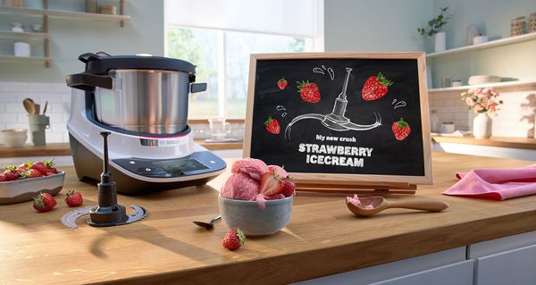 La imagen es la cuchilla trituradora de Cookit con su especial forma ondulada. En el fondo, hay un tazón de helado, una pizarra que dice "Helado de cereza de la Selva Negra" y el robot de cocina Cookit.