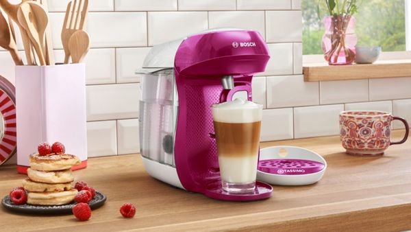 TASSIMO HAPPY in the colour pink brewing a Latte Macchiato on a kitchen worktop and a plate of pancakes next to it.