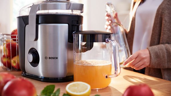 Eine Frau gibt mit einer Hand einen Apfel in den Zentrifugalentsafter VitaJuice von Bosch. Gleichzeitig greift sie mit der anderen Hand nach einem weiteren Apfel, der vor ihr liegt.