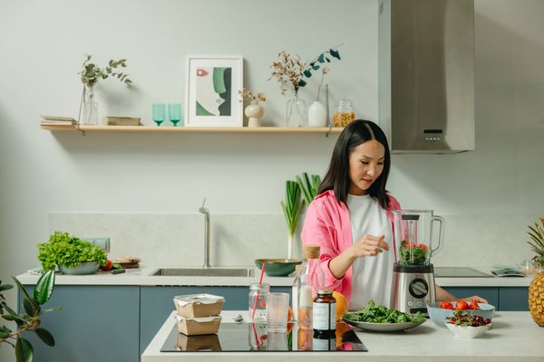 Kobieta w białej koszulce i różowej bluzce w kuchni przygotowuje sok warzywny z porozstawianych warzyw na stole za pomocą blendera.