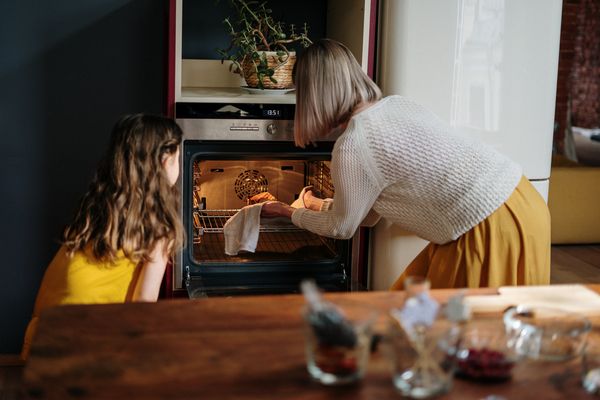 Mama z córką wkładają potrawę do rozgrzanego piekarnika stojącego obok białej lodówki w kuchni.