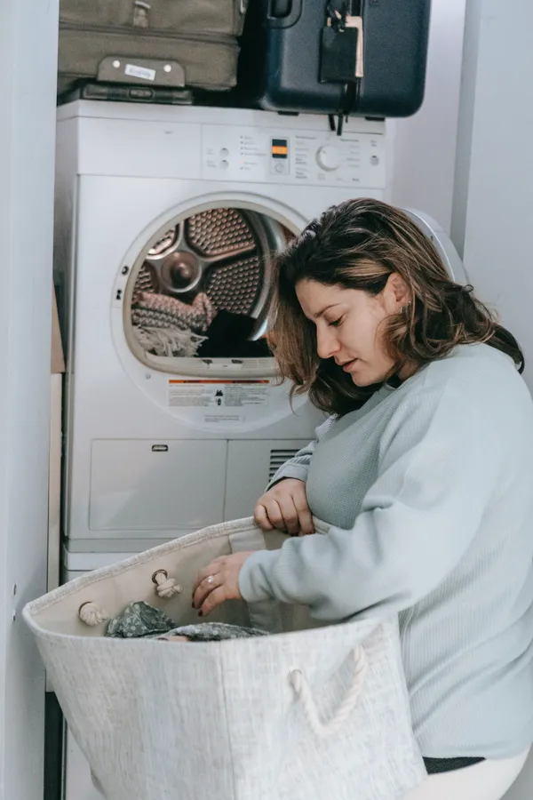 Kobieta w szarej bluzce wkłada pranie do białej pralki/suszarki z beżowej torby na pranie.
