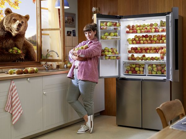 A woman stands infront of a XXL fridge, the doors are open and the fridge is full with apples.