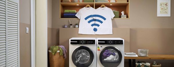 Una moderna lavanderia dispone di due lavatrici, asciugamani colorati e magliette con simbolo Wi-Fi appese sopra uno scaffale di legno.
