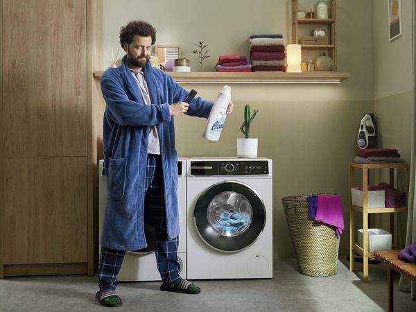 Ein Mann steht in einer Wachküche vor einer Waschmaschine und scannt mit seinem Smartphone das Waschmittel, damit das i-DOS automatische Dosierungssystem die exakte Waschmittel Menge dosiert.