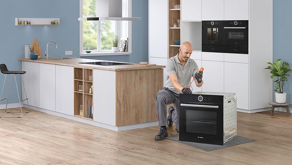 Technician kneels in a white kitchen and repairs a Bosch oven.