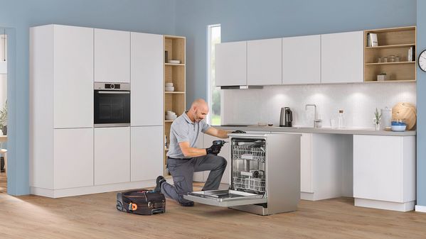 Tekniker på knä i ett vitt kök som reparerar en Bosch-diskmaskin.