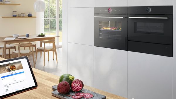 Elegante cocina con horno de la nueva Serie 8 de Bosch con función AirFry y cajón calientaplatos. Bandeja con verduras cocinándose en el horno. Más verduras sobre una tabla de cortar sobre una encimera de cocina.