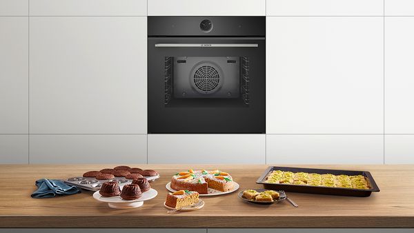 Piekarnik elektryczny Serie|8 marki Bosch w kuchni. Na blacie przed piekarnikiem różne wypieki takie jak ciasto marchewkowe i babeczki.