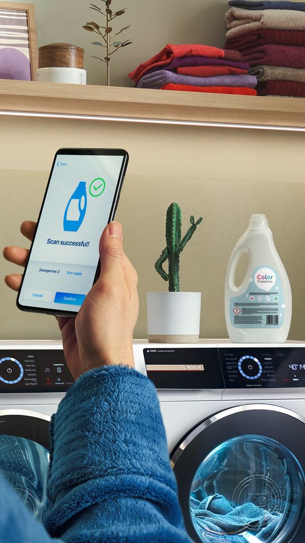 Embalaža tekočega detergenta, ki stoji na pralnih strojih serije 8. Oseba drži pametni telefon, na katerem aplikacija prikazuje, da je bila embalaža detergenta uspešno skenirana.