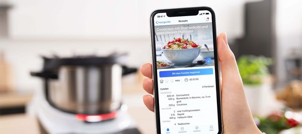 Eine Hand hält ein Smartphone, das ein Rezept für Couscous mit Blumenkohl und Halloumi zeigt, und im Hintergrund sieht man ein Küchengerät.