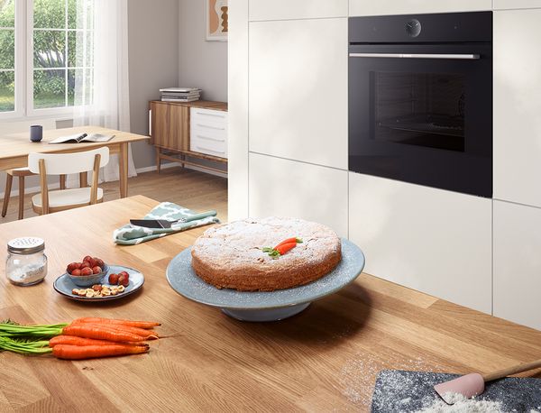 Een levendige worteltaart met decoratief glazuur zit op een blauwe plaat, omringd door verse wortelen in een moderne keuken. 