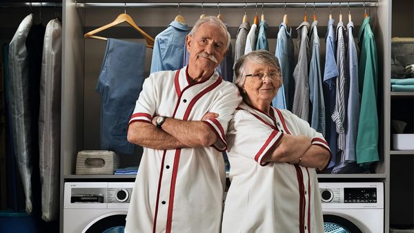 Dwaj wysportowani emeryci ubrani w takie same białe koszulki stoją odwróceni do siebie plecami w eleganckiej pralni.