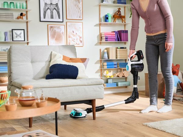 Een vrouw stofzuigt en dweilt de houten vloer in de woonkamer met de Unlimited 7 ProHygienic Aqua. Met zijn flexibele zuigbuis stofzuig je bijzonder makkelijk onder sofa's en tafels.