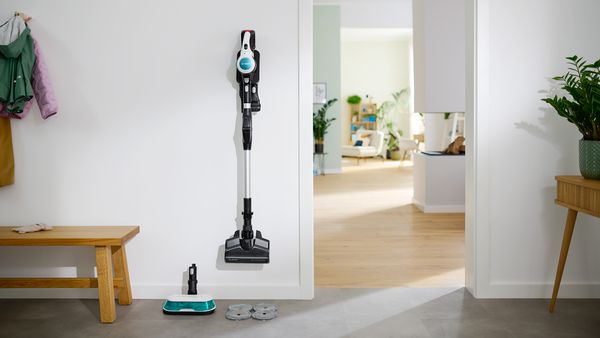 Cordless vacuum cleaner Unlimited 7 Pro Hygienic Aqua hangs in its wall mount in the corridor, with the Aqua nozzle standing on the floor next to it.
