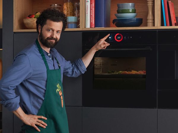 Un homme dans la cuisine, portant un tablier avec un imprimé carotte. Des légumes sont placés à l'intérieur d'un four Bosch Série 8. Son doigt touche l'écran tactile.