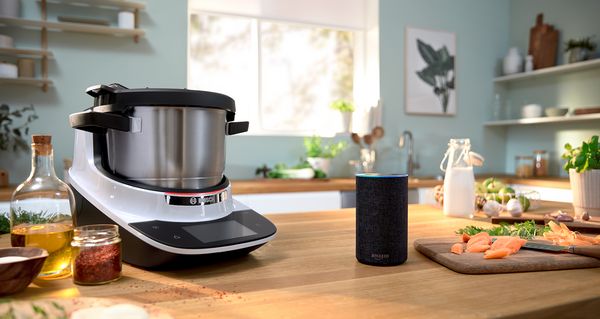 The Cookit stands in the kitchen on a cooking island with a wooden worktop. Next to it is an Alexa and cooking ingredients, such as oil, cream, herbs and a cutting board on which the ingredients are being cut.
