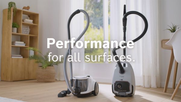 Two Bosch bagless cylinder vacuums stand in front of a window in a study with the words “performance for all surfaces” superimposed over them.