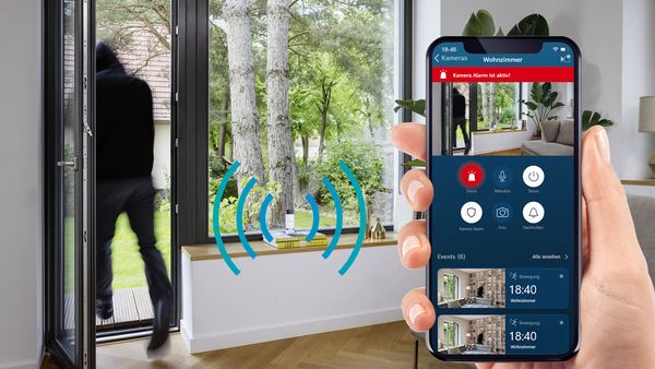 Die Bosch Smart Home 360°-Kamera auf einem Regal und ein Handy mit der Smart Home App, die Liveaufnahmen aus der Wohnung zeigt.