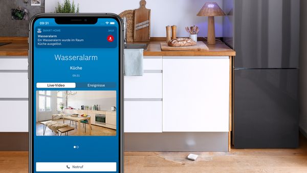 Eine Küche in dem auf dem Boden Wasser ausgelaufen ist und dem Bosch Wassermelder. Im Vordergrund die Smart Home App mit einer Wasseralarmmeldung.